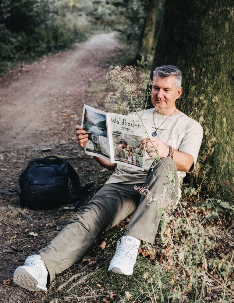 Mann sitzt an einem Baum und liest eine Zeitung, neben ihm liegt ein schwarzer Rucksack.