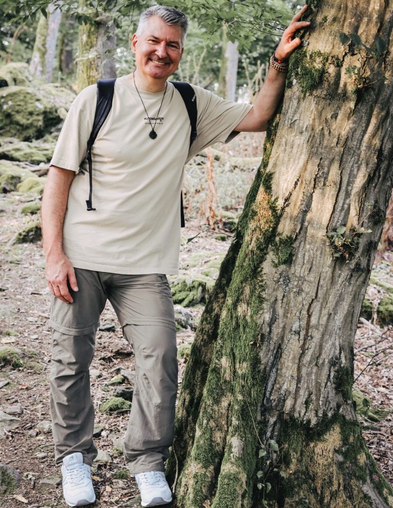 Mann sitzt am Waldweg und liest eine Zeitung, Rucksack neben ihm.
